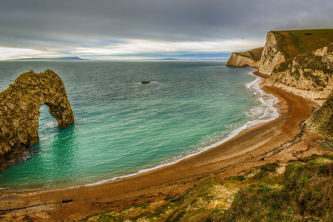 Image of a sandy beach, with green ocean on the left. In the far left a rock formation shaped like an arch is visible. In the far right background there are cliffs.